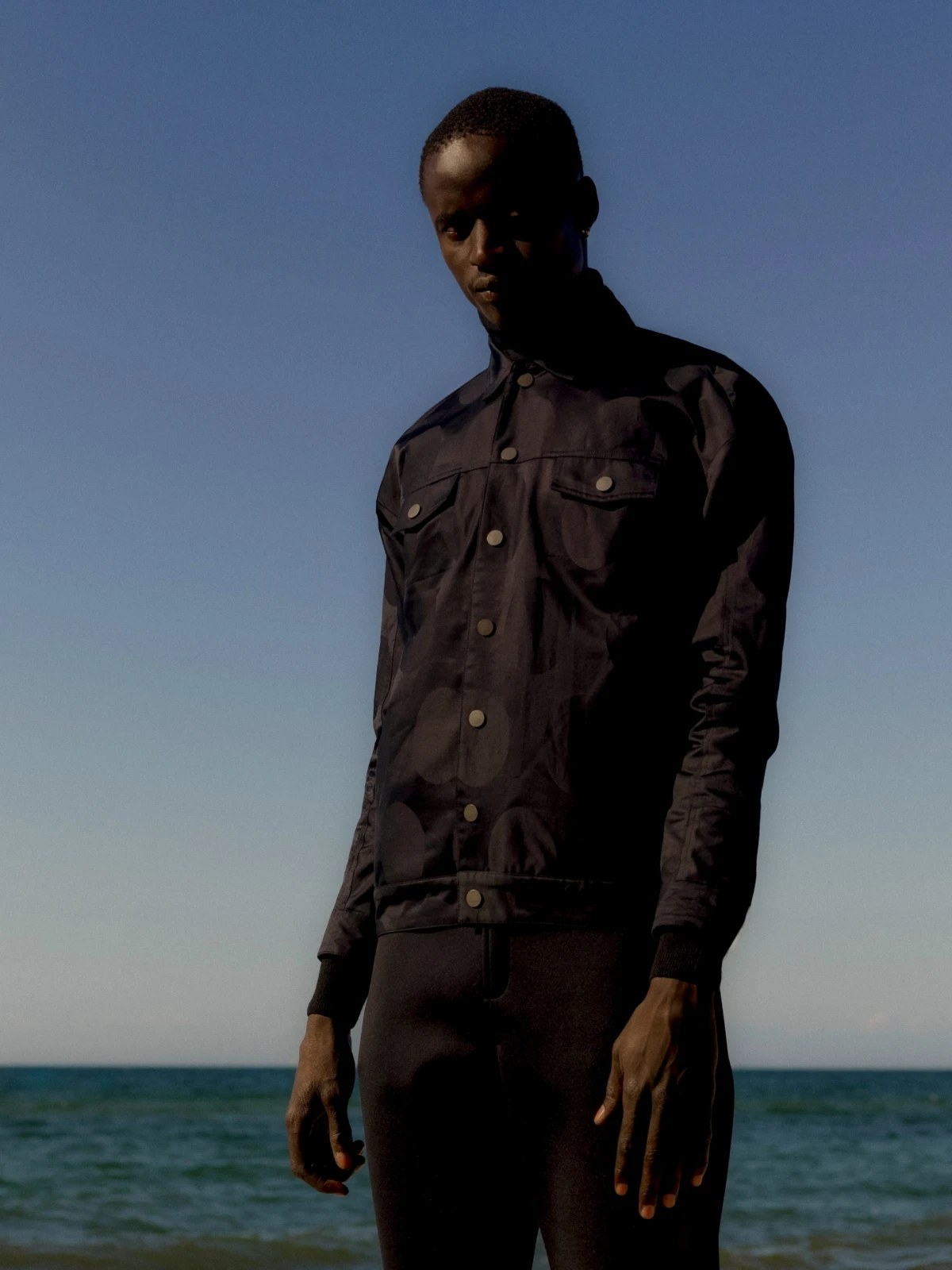 Male model in dark overshirt standing by the sea for Norse Projects fashion campaign